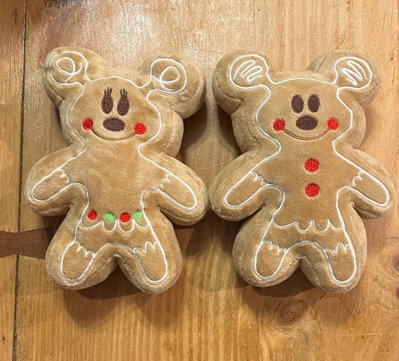 K&ouml;nnte beinhalten: Zwei Pl&uuml;sch-Lebkuchenm&auml;nner in Form von Spielzeugen mit Mickey Mouse-Ohren. Die Spielzeuge sind hellbraun mit wei&szlig;en Verzierungen, roten und gr&uuml;nen Akzenten und braunen Gesichtsz&uuml;gen. Die Spielzeuge liegen auf einer Holzoberfl&auml;che.