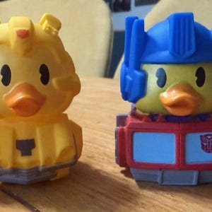 May include: Two yellow rubber duck toys, one resembling Bumblebee and the other Optimus Prime from Transformers. The Bumblebee duck has yellow armor, while the Optimus Prime duck has a red and blue design with the Autobot symbol.