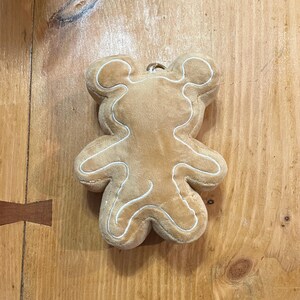 K&ouml;nnte beinhalten: Ein pl&uuml;schiges, hellbraunes B&auml;renornament mit wei&szlig;en N&auml;hten, die seine Form umrei&szlig;en. Der B&auml;r hat oben eine kleine Schlaufe zum Aufh&auml;ngen. Das Ornament besteht aus weichem Material und &auml;hnelt einem Lebkuchen.