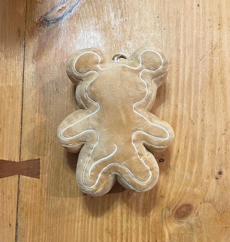 K&ouml;nnte beinhalten: Ein pl&uuml;schiges, hellbraunes B&auml;renornament mit wei&szlig;en N&auml;hten, die seine Form umrei&szlig;en. Der B&auml;r hat oben eine kleine Schlaufe zum Aufh&auml;ngen. Das Ornament besteht aus weichem Material und &auml;hnelt einem Lebkuchen.