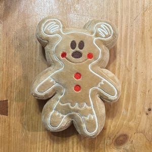 K&ouml;nnte beinhalten: Ein Pl&uuml;sch-Lebkuchenmann in Form einer Maus. Er hat einen braunen K&ouml;rper mit wei&szlig;en Zuckergussdetails und roten Akzenten. Der mausf&ouml;rmige Lebkuchenmann hat ein l&auml;chelndes Gesicht mit braunen Augen und roten Wangen. Er hat zwei rote Kn&ouml;pfe.