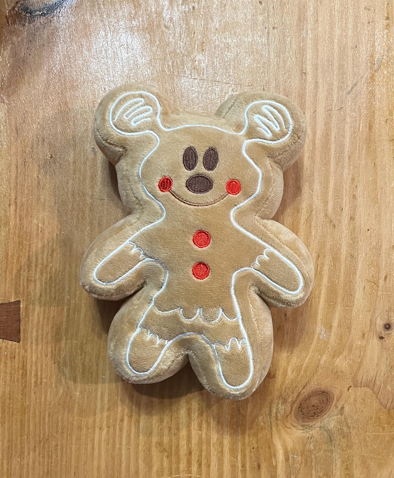 K&ouml;nnte beinhalten: Ein Pl&uuml;sch-Lebkuchenmann in Form einer Maus. Er hat einen braunen K&ouml;rper mit wei&szlig;en Zuckergussdetails und roten Akzenten. Der mausf&ouml;rmige Lebkuchenmann hat ein l&auml;chelndes Gesicht mit braunen Augen und roten Wangen. Er hat zwei rote Kn&ouml;pfe.