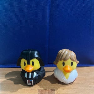 May include: Two rubber ducks styled as characters. One duck features a black helmet, cape, and chest control panel. The other duck has light brown hair and a white robe. Both ducks have yellow faces and orange beaks, standing side by side.