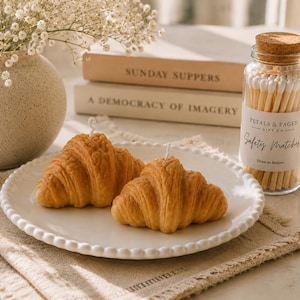 May include: Two golden-brown croissant-shaped candles with visible wicks sit on a white beaded plate. Beside them is a clear glass jar filled with white-tipped safety matches, featuring a label that reads Petals & Pages Gift Co. Safety Matches.