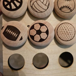 May include: Wooden game pieces featuring sports ball designs: basketball, volleyball, baseball, football, soccer, and golf ball. The pieces are arranged on a light-colored wooden board with three empty circular spaces.