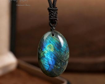 May include: Oval labradorite pendant with iridescent blue and green hues, suspended from a black cord. The stone's surface displays a play of color. The pendant is a piece of handmade jewelry.