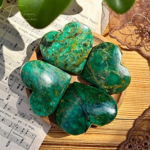 May include: Five heart-shaped green stones with gold veining are arranged on a small wooden plate. The stones have a polished, glossy finish. The background includes sheet music and a decorative lace doily.