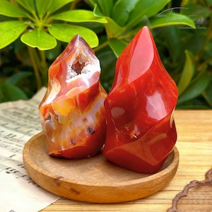 May include: Two polished carnelian flame-shaped crystals. One is a vibrant red, the other is a mix of red, orange, and white, with a cavity. They sit on a small wooden tray, with sheet music in the background.