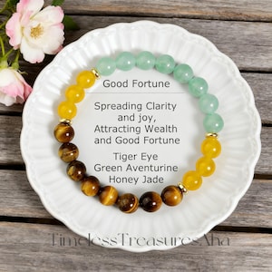 May include: A bracelet featuring tiger eye, green aventurine, and honey jade beads. The bracelet is displayed on a white plate. Text on the plate reads "Good Fortune" and "Timeless Treasures Aha". The beads are yellow, brown, and green.