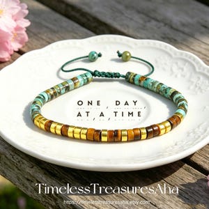 May include: A beaded bracelet with turquoise, brown, and gold-colored beads. The bracelet is on a white plate with the text "ONE DAY AT A TIME". The bracelet is strung on a green cord.