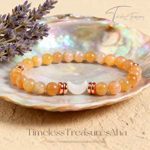 May include: A beaded bracelet featuring orange and peach-colored gemstones, accented with rose gold-colored metal beads and a white crescent moon charm. The bracelet is displayed on a seashell with lavender sprigs. The text "Timeless Treasures Aha" is visible.