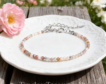 May include: A delicate bracelet with small, colorful beads and silver accents rests on a white, patterned plate. The bracelet features a variety of earth-toned beads, including pink, orange, and brown. The background includes pink roses and greenery.