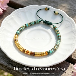 Pode incluir: Uma pulseira de contas com contas turquesa, castanhas e douradas. A pulseira está sobre um prato branco. Possui um cordão verde ajustável. O texto "Timeless TreasuresAha" e um endereço web são visíveis.
