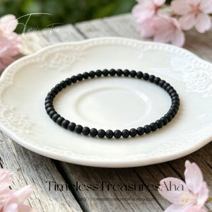 May include: A circular bracelet composed of small, uniform, matte black beads arranged in a continuous loop. The beads have a smooth, non-reflective finish and are strung together to form a simple, minimalist band without any visible clasps or charms.