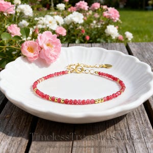 May include: A delicate bracelet featuring small, red gemstone beads interspersed with gold-colored beads. The bracelet is displayed on a white, scalloped-edge plate, with pink roses in the background. The clasp and chain are gold-colored.