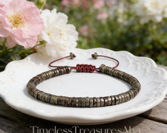 May include: A beaded bracelet with dark, metallic-colored beads. The bracelet is on a white plate with a red cord and knot closure. The background features pink and white flowers and the text "Timeless Treasures Aha".