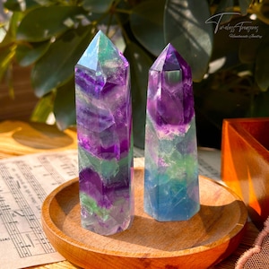 May include: Two fluorite crystal points, one green and purple, the other blue and purple, on a wooden coaster. The crystals are polished and have a pointed tip.