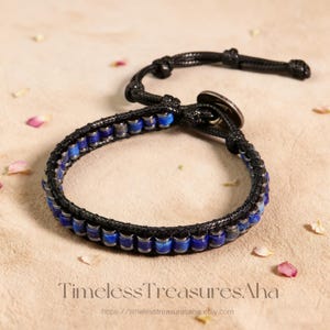 May include: A black leather bracelet with blue and silver beads. The bracelet has a button clasp and a braided design. The words "Timeless Treasures Aha" are at the bottom of the image.