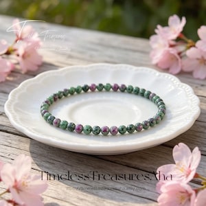 May include: A beaded bracelet featuring green and red gemstones, resting on a white, scalloped-edge plate. The bracelet is made of small, round beads. The background includes pink flowers and the text "Timeless Treasures Aha Handmade Jewelry".