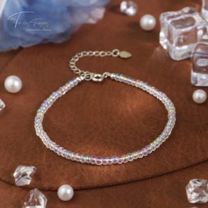 May include: A delicate silver bracelet with a heart charm and iridescent glass beads. The bracelet is on a brown surface with white pearls and clear glass cubes.