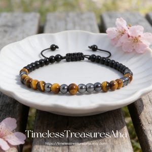 May include: A beaded bracelet featuring a combination of black, brown, and gray beads. The bracelet is displayed on a white plate with pink flowers, and the text "Timeless Treasures Aha" is visible.