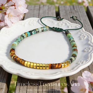 May include: A beaded bracelet featuring a mix of turquoise-colored stone beads with dark veining, brown tiger eye beads, and polished gold-tone metal spacers. The bracelet includes a dark green adjustable cord closure with two small beads.