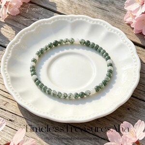 Op de afbeelding: Een delicate kralenarmband met kleine, ronde, mosgroene gespikkelde edelsteenkralen afgewisseld met gladde, ondoorzichtige witte kralen. De armband is in een cirkelvorm gerangschikt op een wit geschulpt keramieken schoteltje.