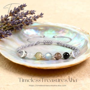 May include: A gray macrame bracelet with a series of colorful gemstone beads, including white, yellow, blue, brown, and black. The bracelet features silver crescent moon charms on each end. The bracelet is displayed on a pearlescent shell.