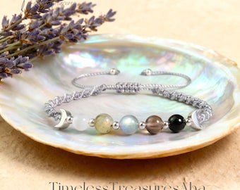 May include: A gray macrame bracelet with a series of colorful gemstone beads, including white, yellow, blue, brown, and black. The bracelet features silver crescent moon charms on each end. The bracelet is displayed on a pearlescent shell.