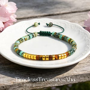 May include: A beaded bracelet with turquoise, brown, and gold-colored beads. The bracelet is on a white plate, with a green cord and a macrame knot. The text "Timeless Treasures Aha" and a website address are at the bottom.