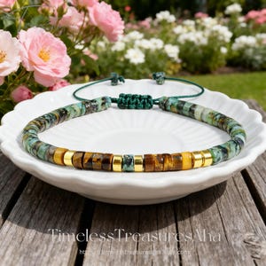Peut inclure: Un bracelet de perles composé de perles turquoise, œil de tigre et dorées. Le bracelet est monté sur un cordon vert avec une fermeture à nœud réglable. Le bracelet est présenté sur une assiette blanche avec un fond floral.