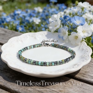 May include: A turquoise and silver beaded bracelet displayed on a white, scalloped-edge plate. The bracelet features small, round turquoise beads interspersed with silver-colored metal beads. The background includes blue and white flowers and the text "Timeless Treasures Aha".