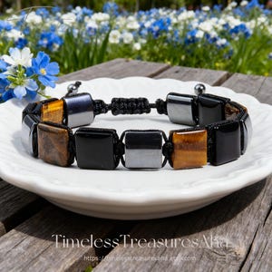 Może przedstawiać: Bransoletka z kwadratowymi koralikami w kolorze czarnym, tygrysiego oka i srebrnym. Bransoletka jest nawleczona na czarny sznurek z regulowanym zapięciem na supeł. Prezentowana jest na białym talerzu z rozmytym kwiatowym tłem. Widoczny jest tekst "Timeless Treasures Aha".