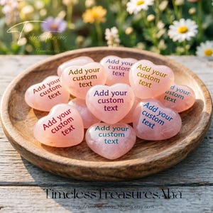 Peut inclure: Un bol en bois contient plusieurs pierres roses en forme de cœur. Chaque pierre porte l'inscription "Add your custom text" en différentes couleurs. L'arrière-plan présente des fleurs et du feuillage flous. Le nom de la boutique est en bas.