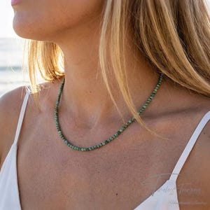 May include: A delicate necklace made of small, round, green beads. The necklace is worn against a person's skin, with a white top visible. The background shows a glimpse of the ocean and sunlight.