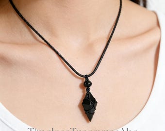 Puede incluir: Collar con colgante de obsidiana negra de forma geométrica, envuelto en cordón negro, suspendido de un cordón trenzado negro. Una pequeña cuenta negra está colocada encima del colgante. El collar se muestra sobre un fondo blanco.