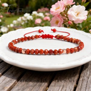 May include: Red beaded bracelet with rose gold accents, displayed on a white plate. The bracelet features faceted beads and a sliding knot closure. The background includes pink flowers and the text "Timeless Treasures".