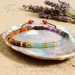 May include: A colorful beaded bracelet with a variety of gemstones, including orange, brown, green, and blue, accented with gold-colored metal spacers. The bracelet is displayed on a white, shell-shaped dish, with lavender sprigs in the background.