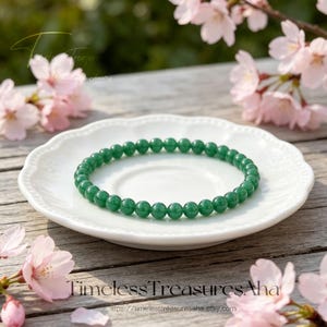 May include: A green beaded bracelet rests on a white, scalloped-edge plate. The bracelet is composed of small, round, polished beads. The plate sits on a wooden surface, with pink flowers in the background. The text "Timeless Treasures Aha" is visible.
