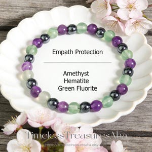 May include: A beaded bracelet featuring amethyst, hematite, and green fluorite stones. The bracelet is composed of round, polished beads in shades of purple, green, and black. Text on the image reads "Empath Protection".