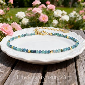 May include: A delicate beaded bracelet with blue-green gemstones and gold accents. The bracelet is displayed on a white plate, with a gold clasp and chain. The background features pink and white flowers and the text "Timeless Treasures Aha".