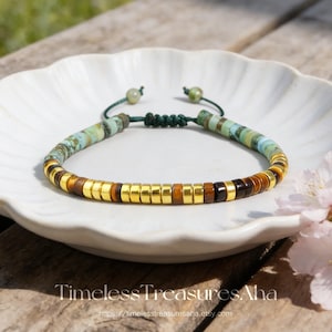 May include: A beaded bracelet featuring a mix of turquoise-colored stone discs, polished gold-tone metal spacers, and brown tiger eye beads. The bracelet is secured with a dark green adjustable cord closure with two small green beads at the ends.