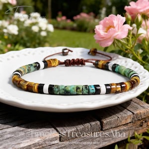 Peut inclure: Un bracelet de perles avec un cordon marron et un nœud réglable. Le bracelet présente un motif de perles noires, blanches, turquoise et œil de tigre. Le bracelet est présenté sur une assiette blanche avec un arrière-plan flou de fleurs.