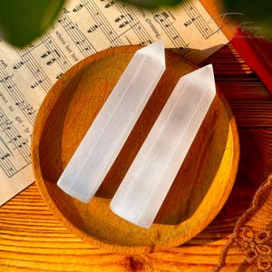 Puede incluir: Dos varitas de selenita blanca sobre un plato de madera. Las varitas son lisas y tienen una textura natural estriada.