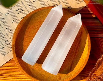 May include: Two white selenite wands on a wooden plate. The wands are smooth and have a natural, striated texture.