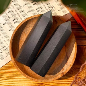 Puede incluir: Dos puntas de cristal de obsidiana negra en un cuenco de madera. Las puntas son lisas y pulidas, con una punta afilada en la parte superior. El cuenco está hecho de madera oscura y tiene un acabado natural.