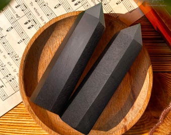 Può includere: Due punte di cristallo di ossidiana nera in una ciotola di legno. Le punte sono lisce e lucidate, con una punta affilata in cima. La ciotola è in legno scuro e ha una finitura naturale.