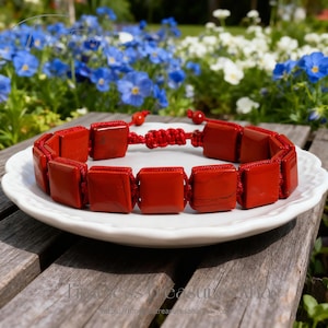 May include: A red square stone bracelet with a macrame knot closure. The bracelet is made of polished, square-cut red stones strung together with red cord. The bracelet is displayed on a white plate.