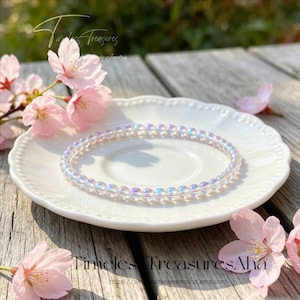 May include: A clear, iridescent beaded bracelet rests on a white, scalloped-edge plate. Pink cherry blossoms and petals are scattered around the plate on a wooden surface. The text "Timeless Treasures Aha" is visible.