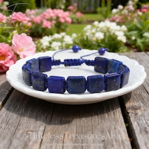 May include: A blue lapis lazuli bracelet with square beads. The bracelet is displayed on a white plate, with a blue macrame closure. The background features pink and white flowers and a wooden surface.